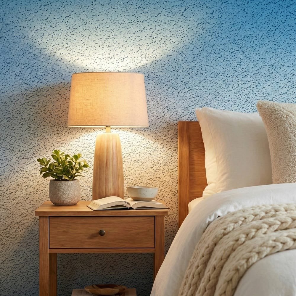 A zoomed-in of sapphire ombre wallpaper in a bedroom with bedside table, lamp, and neutral linens.